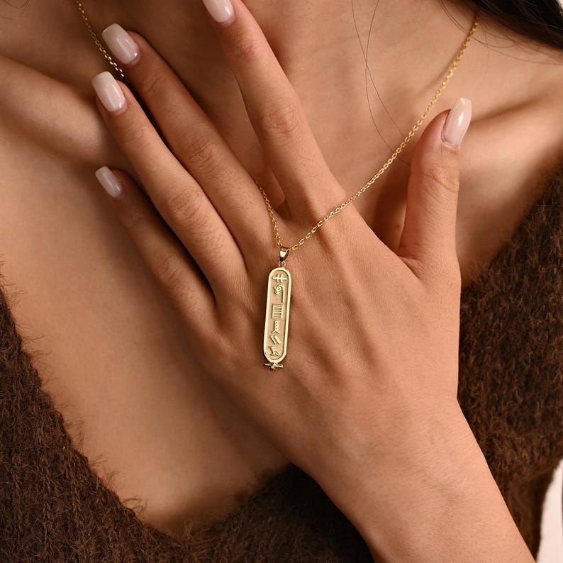 K&ouml;nnte beinhalten: Goldfarbene Anh&auml;ngerkette mit einem l&auml;nglichen rechteckigen Anh&auml;nger. Der Anh&auml;nger zeigt eingravierte Hieroglyphen-Symbole. Die Halskette wird vor einem braunen Hintergrund pr&auml;sentiert.