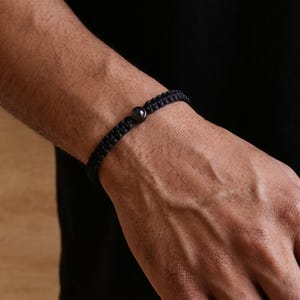 Peut inclure: Bracelet noir tress&eacute; avec une seule perle sombre et ronde. Le bracelet est port&eacute; au poignet, mettant en valeur la texture tress&eacute;e et le contraste de la perle sombre sur la peau.