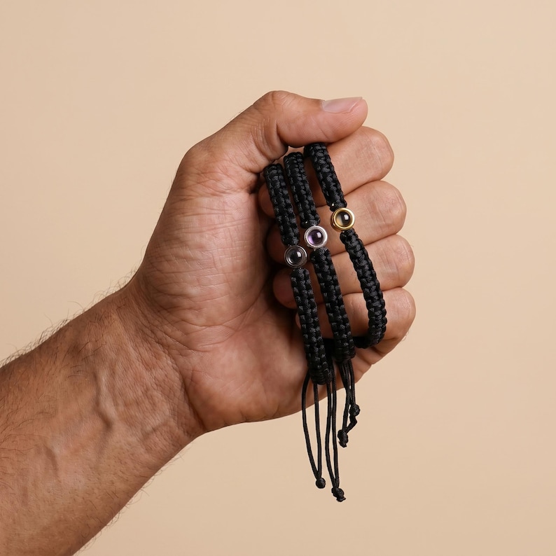 Peut inclure: Trois bracelets noirs tress&eacute;s avec des liens r&eacute;glables. Chaque bracelet est orn&eacute; d'une petite pierre pr&eacute;cieuse ronde dans un sertissage dor&eacute; ou argent&eacute;. Les pierres pr&eacute;cieuses sont violettes et dor&eacute;es. Les bracelets sont tenus dans une main sur fond neutre.