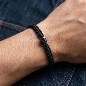 Peut inclure: Bracelet tress&eacute; noir avec une pierre pr&eacute;cieuse violet fonc&eacute; au centre. Le bracelet est fait d'un mat&eacute;riau tiss&eacute; et pr&eacute;sente un design simple et &eacute;l&eacute;gant. La pierre pr&eacute;cieuse est ronde et sertie dans un sertissage en m&eacute;tal fonc&eacute;.