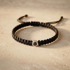 Peut inclure: Un bracelet noir tress&eacute; avec une petite perle ronde de couleur argent&eacute;e. Le bracelet est fait d'un mat&eacute;riau &eacute;pais et tiss&eacute; et est r&eacute;glable. Le fond est une surface neutre de couleur claire.