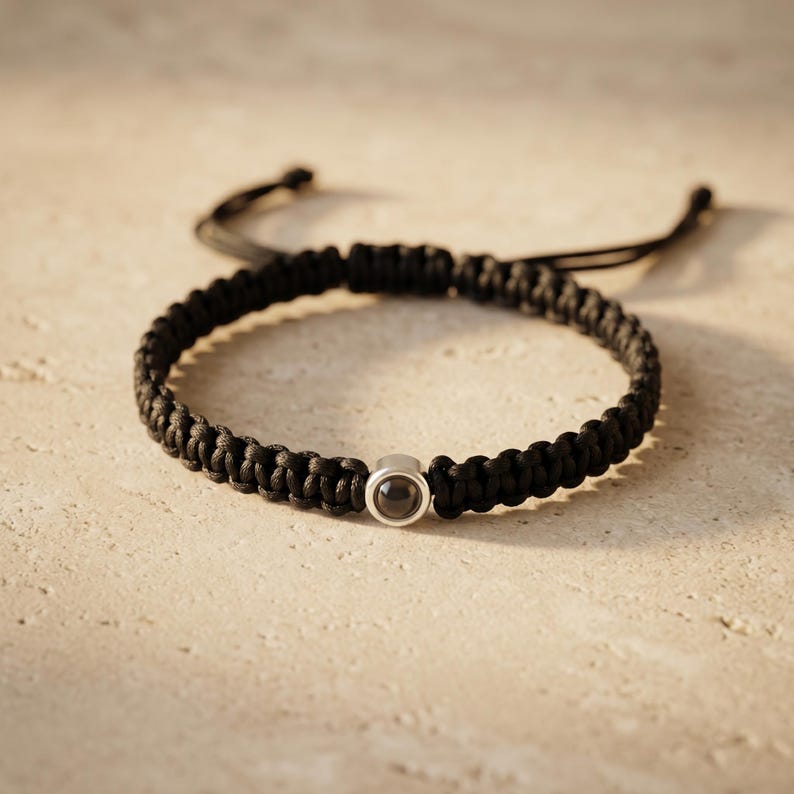 Peut inclure: Un bracelet noir tress&eacute; avec une petite perle ronde de couleur argent&eacute;e. Le bracelet est fait d'un mat&eacute;riau &eacute;pais et tiss&eacute; et est r&eacute;glable. Le fond est une surface neutre de couleur claire.