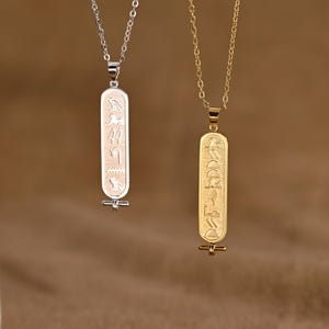 K&ouml;nnte beinhalten: Zwei Anh&auml;nger-Halsketten mit &auml;gyptischem Hieroglyphen-Design. Eine Halskette ist silberfarben, die andere goldfarben. Die rechteckigen Anh&auml;nger haben eingravierte Symbole und h&auml;ngen an filigranen Ketten. Der Hintergrund ist warmbraun.