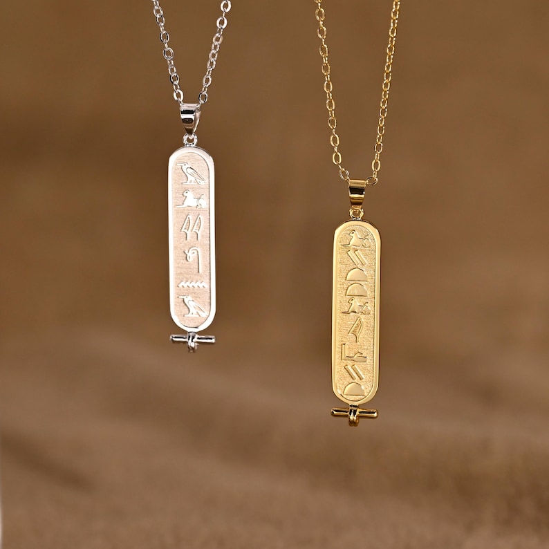 K&ouml;nnte beinhalten: Zwei Anh&auml;nger-Halsketten mit &auml;gyptischem Hieroglyphen-Design. Eine Halskette ist silberfarben, die andere goldfarben. Die rechteckigen Anh&auml;nger haben eingravierte Symbole und h&auml;ngen an filigranen Ketten. Der Hintergrund ist warmbraun.