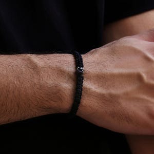 Peut inclure: Bracelet tress&eacute; noir avec une petite pierre pr&eacute;cieuse bleu fonc&eacute; sertie dans un m&eacute;tal argent&eacute;. Le bracelet est port&eacute; au poignet, mettant en valeur la texture tress&eacute;e et le contraste de la pierre sombre sur la peau.