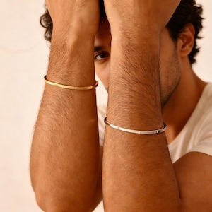 Könnte beinhalten: Ein goldfarbenes und ein silberfarbenes Armreif-Armband. Das goldfarbene Armband befindet sich am linken Arm, das silberfarbene am rechten Arm. Beide Armbänder haben ein schlichtes, minimalistisches Design.