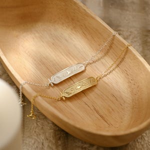 K&ouml;nnte beinhalten: Zwei zarte Halsketten mit rechteckigen Anh&auml;ngern mit Hieroglyphen-Designs. Eine Kette ist silberfarben, die andere goldfarben. Die Halsketten liegen in einer Holzschale und zeigen ihre filigranen Details und ihr elegantes Design.