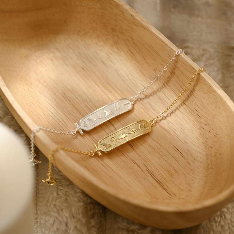 K&ouml;nnte beinhalten: Zwei zarte Halsketten mit rechteckigen Anh&auml;ngern mit Hieroglyphen-Designs. Eine Kette ist silberfarben, die andere goldfarben. Die Halsketten liegen in einer Holzschale und zeigen ihre filigranen Details und ihr elegantes Design.