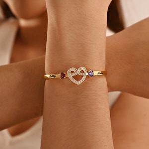 Puede incluir: Pulsera dorada con un centro en forma de corazón incrustado con pequeñas piedras transparentes. La pulsera tiene un diseño delicado con una gema roja y otra púrpura a cada lado del corazón. Los nombres "Ashley" y "Laura" están grabados.