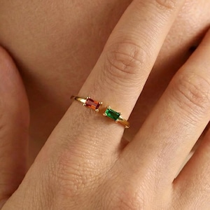 May include: A gold ring featuring two rectangular gemstones, one red and one green. The ring is worn on a finger, with the gemstones positioned side-by-side. The ring's design is minimalist and elegant, suitable for everyday wear.