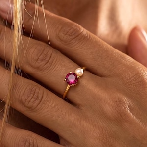 May include: A gold ring featuring a round, vibrant pink gemstone set next to a small, white, spherical pearl. The band is thin and polished, showcasing a minimalist design that highlights the contrast between the bright pink stone and the pearl.