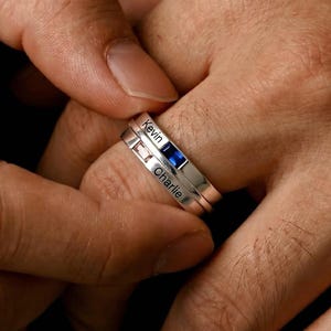 Peut inclure: Deux bagues en argent, l'une avec une pierre précieuse bleue et l'autre avec le nom "Kevin" gravé. La deuxième bague porte le nom "Charlie". Les bagues sont sur le doigt d'une personne, avec un fond sombre.