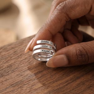 May include: A silver-colored wrap ring featuring three stacked bands. Each band is engraved with a name in a black sans-serif font. The names visible on the bands are Goldie, John, and Kayla, arranged in a vertical sequence on the metal surface.