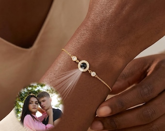 May include: A gold-toned chain bracelet featuring a central circular pendant with a dark stone. The pendant projects a circular light beam displaying a color photograph of a woman in a pink shirt and a man in a dark shirt standing together outdoors.