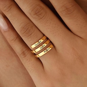 May include: Three gold-colored rings stacked on a finger, each engraved with a name: Goldie, John, and Kayla. The rings are simple bands with a polished finish, creating a minimalist jewelry aesthetic.