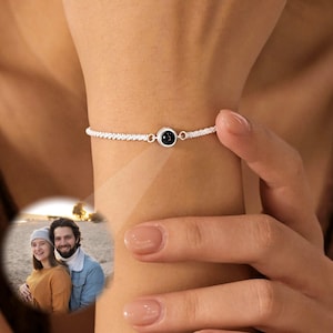 May include: A silver-toned chain bracelet featuring a small, circular central charm. The charm contains a projection lens that displays a circular, full-color photograph of two people standing together outdoors on a beach or sandy surface.