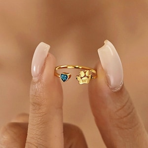 Op de afbeelding: Een gouden open ring met een blauwe hartvormige edelsteen en een pootafdrukontwerp. De ring wordt door de vingers van een persoon omhoog gehouden, waardoor het delicate ontwerp en de details worden benadrukt.