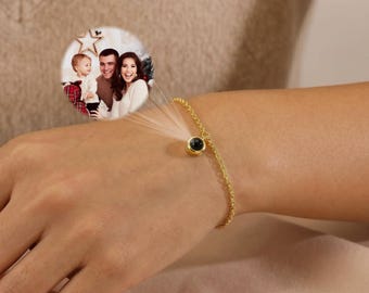May include: A gold-toned chain bracelet featuring a small circular charm that projects a circular image of a family of three. The charm is attached to a delicate link chain that wraps around the wrist, displaying a clear photograph of a child, a man, and a woman.