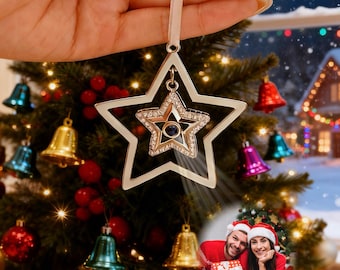 May include: A silver star-shaped Christmas ornament with a smaller, jeweled star inside, hanging from a white ribbon. The ornament is held in front of a decorated Christmas tree with colorful bells and lights. A couple in Santa hats is in the background.