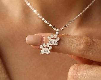 May include: A silver-toned necklace featuring two connected paw print charms. Each charm is engraved with a name, specifically the names Emily and Bear, suspended from a delicate silver-toned chain that rests against the neck.