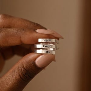 May include: A silver-coloured wrap ring featuring three stacked bands. Each band is engraved with a name in a black sans-serif font. The top band reads Sophie, the middle band reads Evelyn, and the bottom band reads Toby. The metal has a smooth finish.