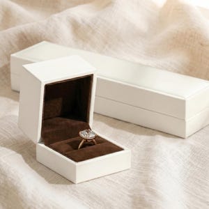 May include: A white ring box with a brown interior, open to display a gold ring with a large, square-cut clear gemstone. Another white box is in the background, both resting on a textured, off-white surface.