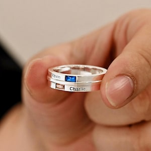 Può includere: Un anello d'argento a due fasce, con i nomi "Kevin" e "Charlie". L'anello ha una pietra blu e una marrone chiaro. L'anello è tenuto nella mano di una persona.