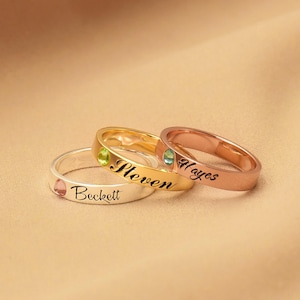 Könnte beinhalten: Drei personalisierte Ringe mit Edelsteinen. Die Ringe sind übereinander gestapelt. Der oberste Ring ist roségoldfarben mit dem eingravierten Namen "Hayes". Der mittlere Ring ist golden mit dem eingravierten Namen "Steven". Der unterste Ring ist silberfarben mit dem eingravierten Namen "Beckett".