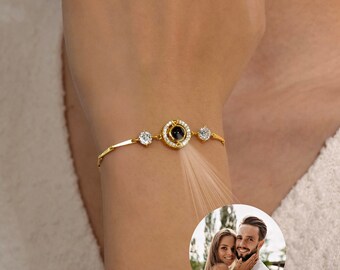 Könnte beinhalten: Ein goldenes Armband mit einem runden, schwarzen Stein in der Mitte, umgeben von kleineren, klaren Steinen. Der Stein projiziert ein holografisches Bild eines Paares.