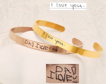 May include: Two gold bracelets with handwritten messages. The top bracelet says "I love you." The bottom bracelet says "Dad I love you 12/12".