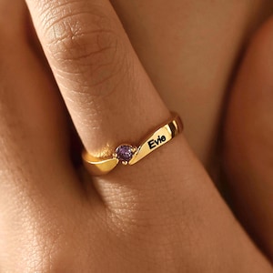 May include: A gold-toned ring featuring a bypass design with a single small round purple gemstone and the name Evie engraved in black lettering on the band. The ring has a smooth, polished finish and a curved shape that wraps around the finger.