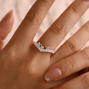 May include: A silver-toned chevron ring featuring a row of small clear stones along the band and a cluster of five multi-colored gemstones at the center, including green, yellow, red, and pink stones arranged in a V-shaped crown design.