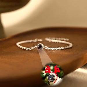Könnte beinhalten: Ein silberfarbenes Kettenarmband mit einem zentralen runden Anhänger, der ein kleines, kreisförmiges Bild eines Mopses mit roten Rentiergeweihen projiziert. Das Armband hat ein feines, strukturiertes Gliederkettendesign und einen Verschluss.