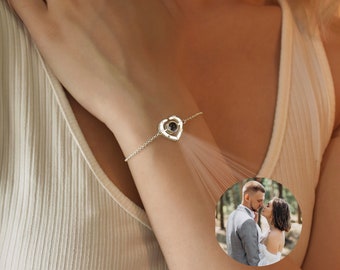 Könnte beinhalten: Ein silberfarbenes Kettenarmband mit einem herzförmigen Anhänger in der Mitte. Der Anhänger enthält eine kreisförmige Projektionslinse, die ein kleines, rundes Farbfoto eines Paares zeigt, das eng beieinander in einem Wald steht.