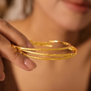 Puede incluir: Tres brazaletes dorados con texto grabado. Los brazaletes son finos y tienen una textura martillada. El texto incluye nombres y fechas. Los brazaletes se sostienen en una mano.