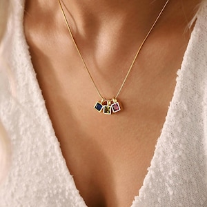 May include: A gold necklace with a fine chain. It has three square pendants, each with a different coloured gemstone: blue, green, and pink. The pendants are set in gold frames, offering a simple, elegant design.