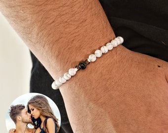 May include: A white marble beaded bracelet with a black bead accent. The bracelet is on a person's wrist.