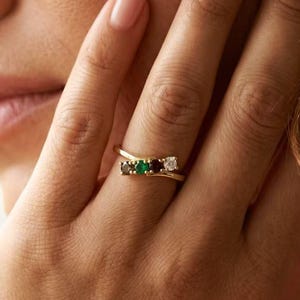 Puede incluir: Un anillo de oro con un diseño de banda cruzada engastado con cuatro pequeñas piedras redondas en fila, incluyendo una piedra blanca clara, una piedra oscura, una piedra verde vibrante y una piedra oscura final, en un engaste de metal.