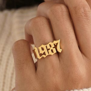 Può includere: Un anello color oro con il numero "1987" in un carattere gotico. L'anello è indossato su un dito. Lo sfondo è di un colore morbido e neutro.