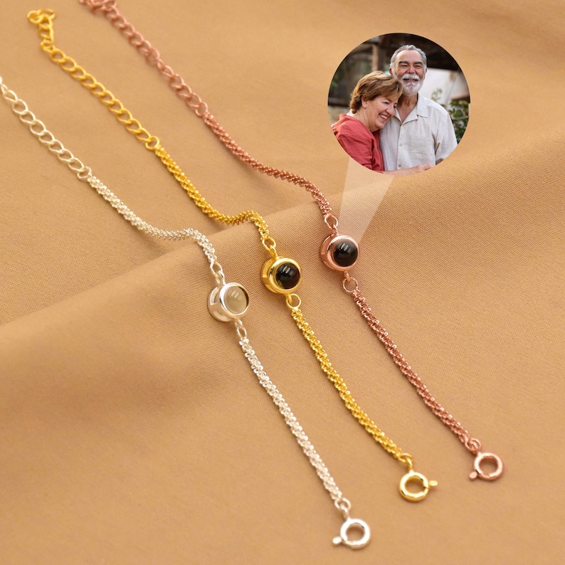 May include: Three delicate chain bracelets in silver, gold, and rose gold tones, each featuring a small circular pendant. One pendant displays a circular inset photograph of a smiling couple. The bracelets include thin metal links and a circular clasp.