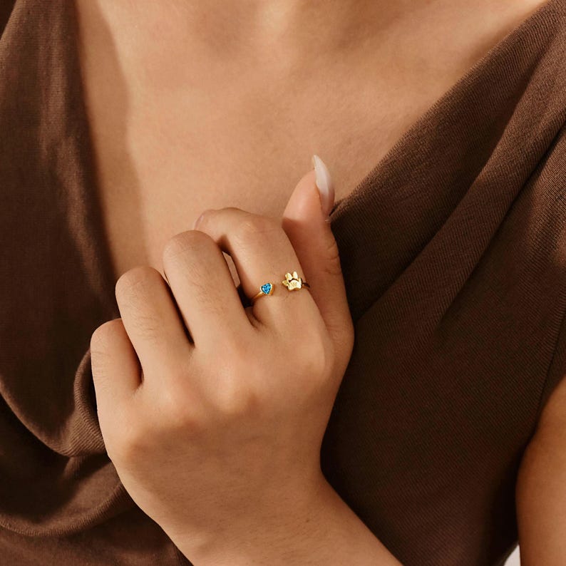 May include: A gold-toned open ring featuring a small blue heart-shaped stone on one end and a detailed animal paw print charm on the other. The band is thin and polished, designed to wrap slightly around the finger with an adjustable open gap.