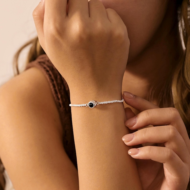 May include: A delicate silver-toned chain bracelet featuring a single, small, circular charm with a smooth, polished black stone center. The bracelet has a fine, textured link design that wraps around the wrist, creating a simple and refined appearance.