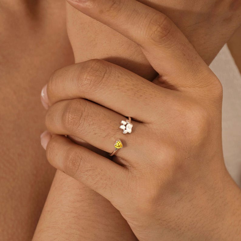 May include: A silver open-style ring featuring a small paw print charm on one end and a single yellow heart-shaped gemstone on the other. The band is thin and metallic, designed to wrap partially around the finger with an adjustable open gap.