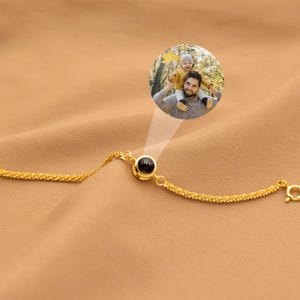 May include: A gold-toned chain bracelet featuring a central circular charm. The charm contains a small, dark, round projection lens that displays a circular color photograph of an adult holding a small child on their shoulders outdoors.