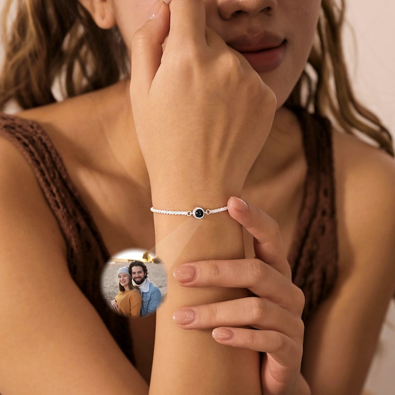 May include: A silver-toned chain bracelet featuring a small, circular central charm. The charm contains a projection lens that displays a circular, full-color photograph of two people standing together outdoors on a beach or sandy surface.