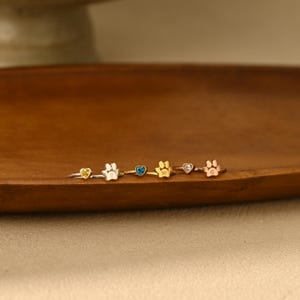 May include: Three rings featuring a heart-shaped gemstone and a paw print charm. The first ring is silver with a yellow stone and the letter E. The second is gold with a blue stone and the letter W. The third is rose gold with a clear stone and the letter G.