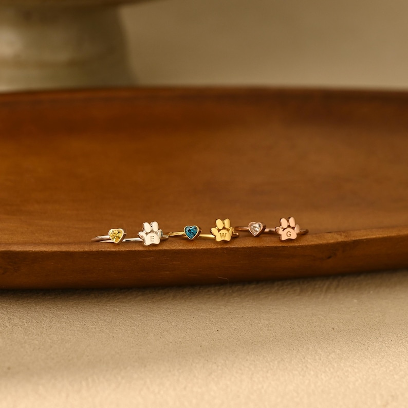May include: Three rings featuring a heart-shaped gemstone and a paw print charm. The first ring is silver with a yellow stone and the letter E. The second is gold with a blue stone and the letter W. The third is rose gold with a clear stone and the letter G.