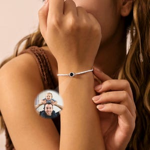 May include: A silver-toned chain bracelet featuring a small, circular central charm. The charm contains a projection lens that displays a color photograph of a person carrying a small child on their shoulders, visible as a circular projection.