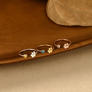 May include: Three open-style rings featuring a small heart-shaped gemstone on one end and a paw print charm on the other. The silver-tone ring has a yellow stone, the gold-tone ring has a blue stone, and the rose gold-tone ring has a clear stone.