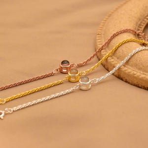 May include: Three delicate chain bracelets in rose gold, yellow gold, and silver tones. Each bracelet features a single small, round, bezel-set charm with a smooth, translucent center stone, connected to a fine, textured link chain design.
